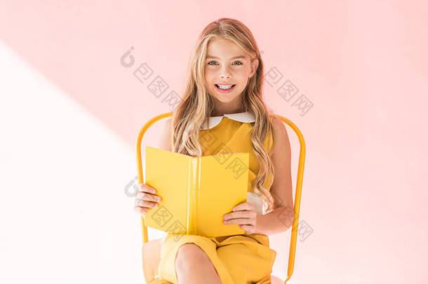 微笑时尚的孩子看书, 而坐在黄色的椅子上粉红色