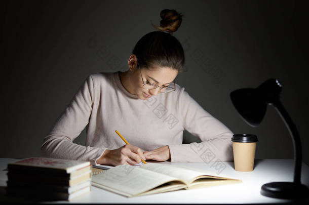 忙自由职业者重写 infrormation 到记事本, 准备文章出版, 读书, 写一些笔记在组织者, 饮料外卖咖啡, 坐在黑暗中, 穿着圆大眼镜
