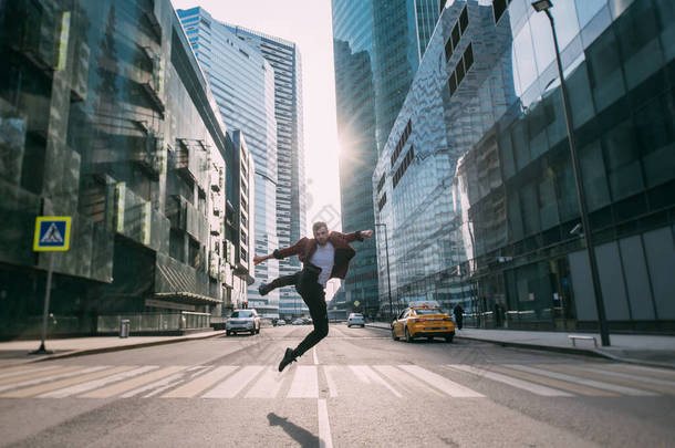 一个男人从市中心的一个人行横道上跳过.在一座大城市的摩天大楼的背景下，一个年轻时髦的小伙子在日落前摆姿势
