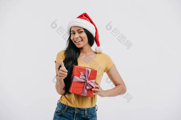 微笑非洲裔美国女孩在圣诞老人的帽子举行目前和显示拇指上孤立的白色