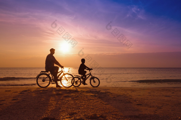 父亲和儿子在海滩上落日下，骑自行车的人的家庭剪影 