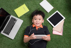可爱的小男孩听音乐在草地上与他的笔记本电脑, 平板电脑和笔记本电脑周围.