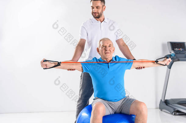 康复治疗师在灰色背景下帮助老年人在健身球上锻炼橡胶胶带
