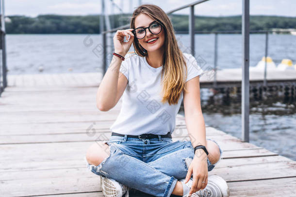 一位留着长发、戴着时髦眼镜的年轻女子躺在湖边的一个木制码头上。穿着牛仔裤和T恤的女孩笑着看着摄像机.