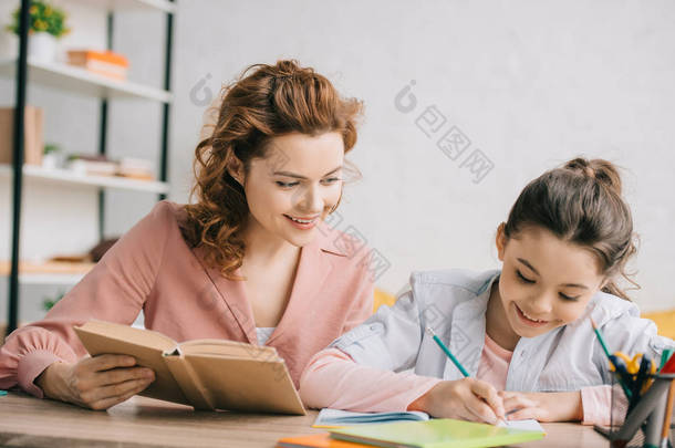美丽的女人拿着书和可爱的女儿写在笔记本上，而做家庭作业一起 