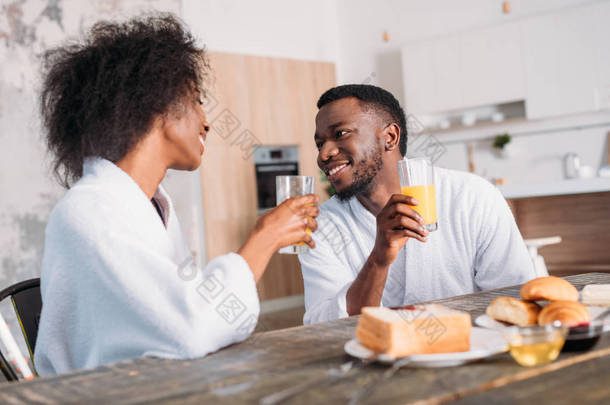 年轻夫妇拿着杯子喝果汁, 坐在餐桌上敬酒和牛角面包 