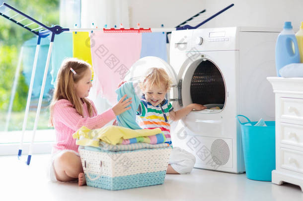 儿童在洗衣房内设有洗衣机或滚筒烘干机。孩子们帮助做家事。现代家庭设备和洗涤洗涤剂在白色阳光的家。烘干机架清洗洗衣服. 