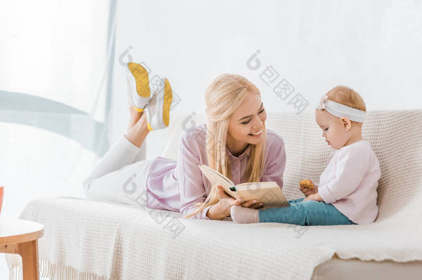 年轻快乐的母亲躺在沙发上<strong>看书</strong>给小女儿, 而幼儿拿着饼干