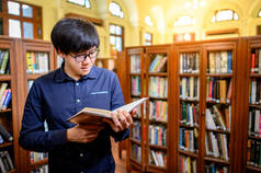 聪明的亚洲人大学生戴着眼镜，坐在老式书架上看书。高校图书馆的教材资源用于教育科目和研究.教育机会奖学金.