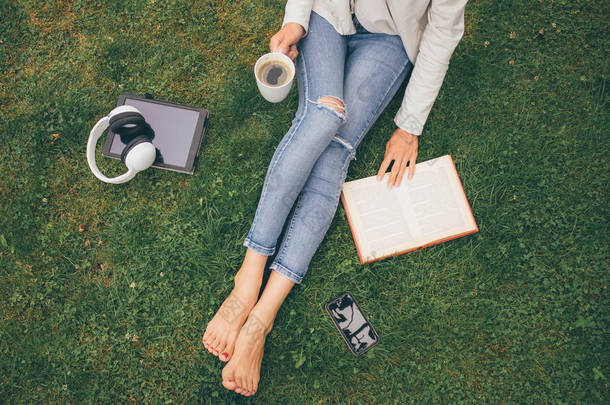 在绿色草地上的妇女坐在公园 (花园) 的最高的看法与智能手机, 耳机, 片剂, 书和咖啡在手里