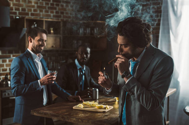 英俊的年轻商人抽雪茄, 而微笑的朋友喝酒精饮料背后 