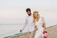 英俊的新郎拥抱美丽的新娘与婚礼花束, 他们走在海滩上