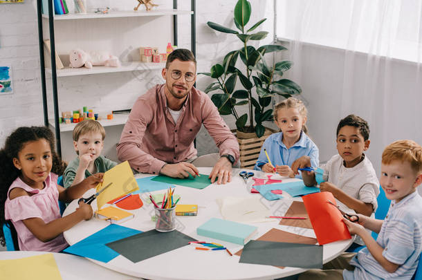 男教师和多种族学龄前儿童坐在桌子上, 在教室里放着五颜六色的纸