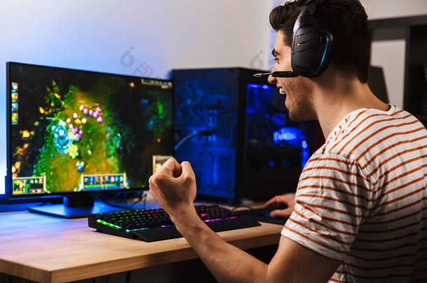 高兴的年轻人的肖像在电脑上玩电子游戏戴着耳机, 并使用背光彩色键盘