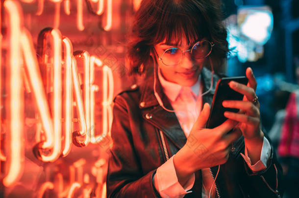 沉思的时髦女孩在眼镜看视频的智能手机满意的互联网连接休息在夜间城市, 迷人的年轻女子阅读信息在网络上的移动聊天在户外