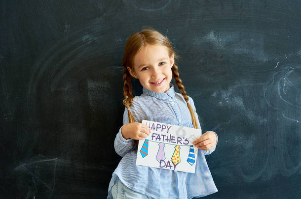 微笑的小女孩的肖像拿着手工<strong>贺卡</strong>为父亲一天摆在学校黑板和看相机
