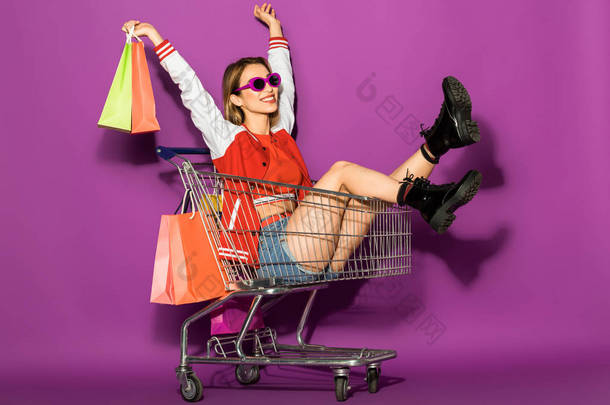美丽快乐的年轻女子在太阳镜持有纸袋, 坐在购物车上的紫罗兰色 