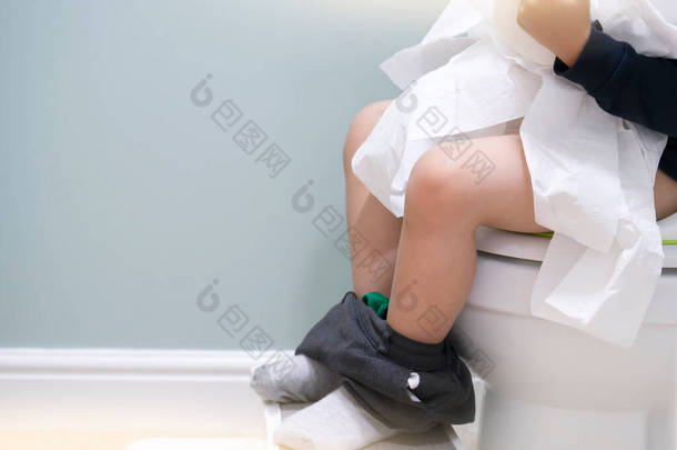 学童坐在马桶上玩卫生纸腿上挂着灰色长裤袜子松软小孩撕破纸巾复制空间训练小孩或健康护理的概念.