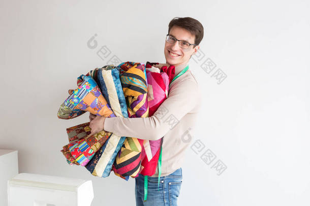 英俊的年轻男裁缝在白色背景上拿着一叠折叠的颜色拼布绗缝被。许多彩色毛毯样式拼凑而成的男人微笑着，手工手中.