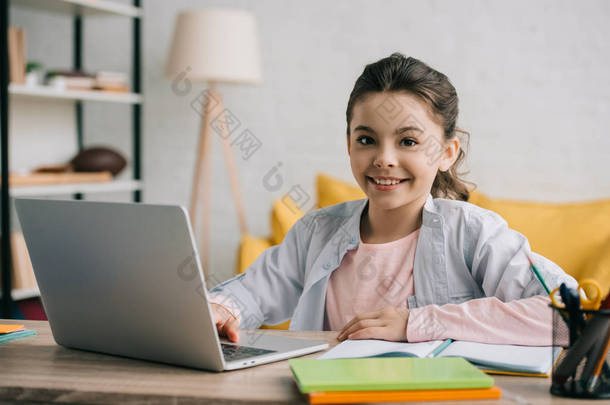 快乐的孩子坐在办公桌前看着相机，在家里用笔记本电脑