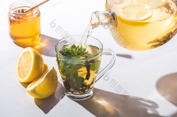 从茶壶倒入柠檬和薄荷的健康茶到杯上白色桌面