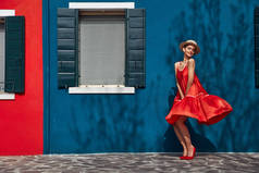 穿着红色连衣裙和高跟鞋的快乐年轻女子头上戴着草帽, 在布拉诺岛上摆着蓝色和红色的房子。墙上有树荫