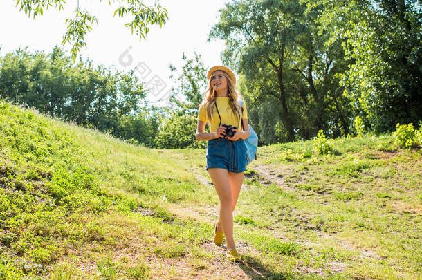年轻微笑的妇女在帽子与相片照相机漫步在夏天公园