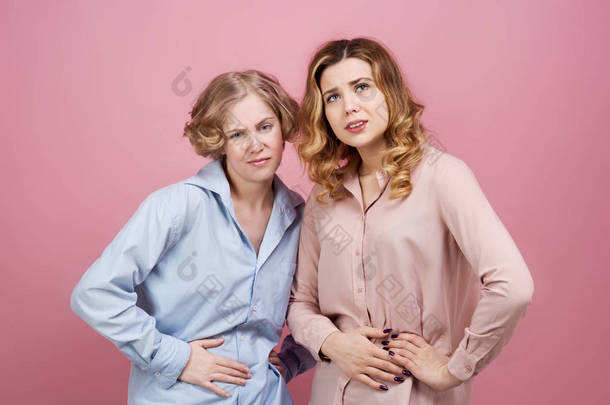 两个年轻的女人用痛苦的表情抱着他们的肚子。工作室<strong>肖像</strong>粉红色背景。胃病、月经痛、消化不良的概念.