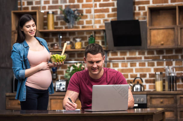 微笑的年轻人使用笔记本电脑和笔记, 而怀孕的妻子拿着蔬菜沙拉玻璃碗 