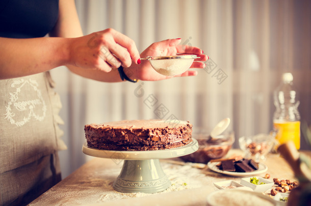 家庭主妇穿着围裙做生日甜点<strong>巧克力蛋糕</strong>的装饰。女人做自制<strong>蛋糕</strong>与简单的食谱，洒糖粉在上面