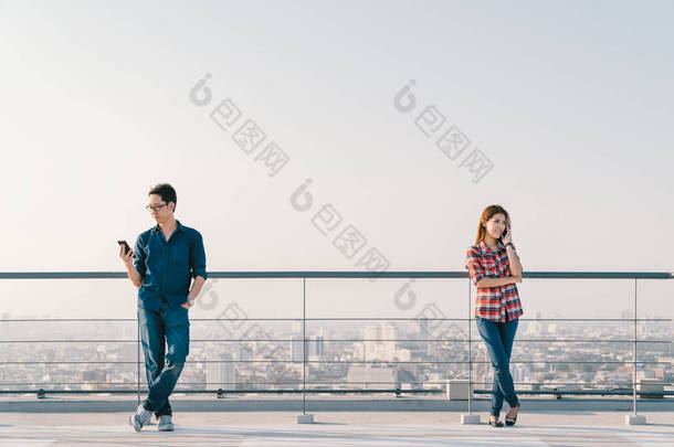 年轻的亚洲夫妇在建筑屋面一起使用电话和智能手机。移动手机设备或信息技术通信的概念。城市景观视图背景，天空副本空间
