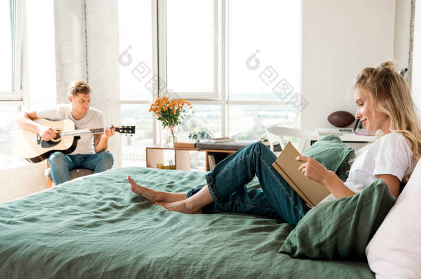年轻的妇女在床上看书, 而男友在家里玩声学吉他