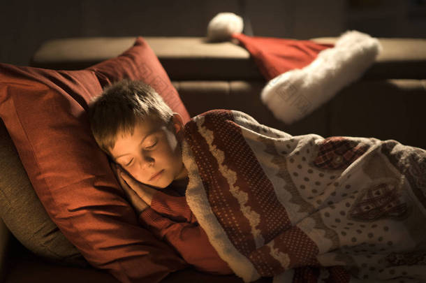 <strong>可爱</strong>的小男孩睡在沙发上的圣诞前夜, 圣诞老人把他的帽子放在他旁边