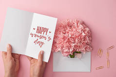 妇女拿着贺卡与愉快的妇女天的文字在康乃馨花附近的裁剪视图在信封在粉红色的背景