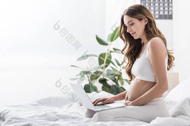 坐在床上使用笔记本电脑的迷人孕妇