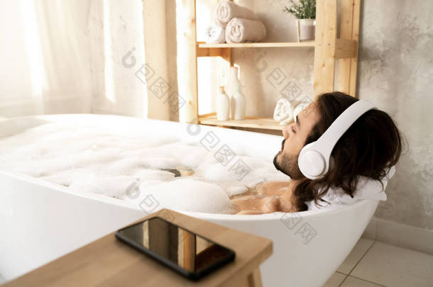 <strong>年轻男子</strong>头戴耳机，躺在布满热水和泡沫的白色浴缸里，享受着悠闲的音乐