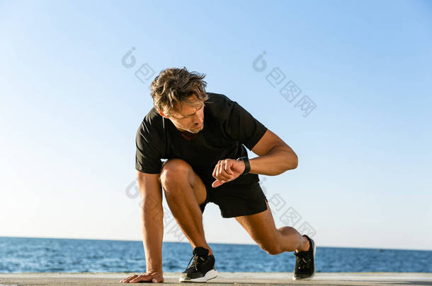 英俊的成人短跑运动员看健身跟踪, 而站在开始的位置运行在海边