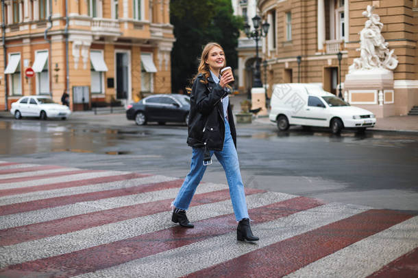 年轻的微笑的妇女在皮革夹克和牛仔裤快乐地看着相机捧着杯咖啡走在舒适的城市街道上行走