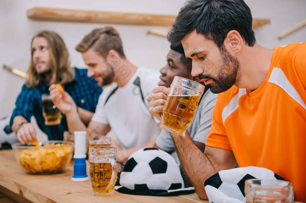 沮丧的年轻人在橙色 t恤的侧面观看啤酒, 而他的朋友坐在酒吧柜台与球帽, 薯条, 风扇喇叭和啤酒在观看足球比赛 
