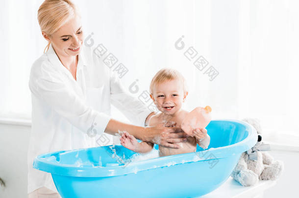 快乐的母亲洗欢快的幼儿儿子拿着瓶子与洗发水 