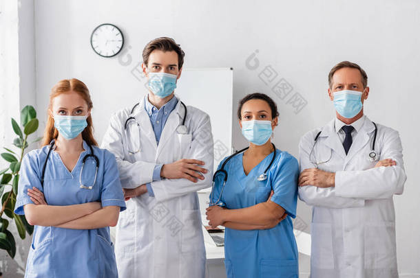 多文化<strong>医生</strong>和<strong>戴口罩</strong>的护士，双手交叉，站在医院里看着相机