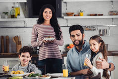 快乐的拉丁母亲在午餐时拿着盘子和食物靠近可爱的儿子、丈夫和女儿