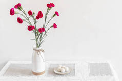 陶瓷花瓶里的桌子上有红色的康乃馨，旁边是一盘糖果。白色背景斯堪的纳维亚风格前视图，复制空间.