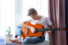 一个英俊的家伙坐在自家房间的窗台上弹着吉他。做音乐的男孩，弹吉他