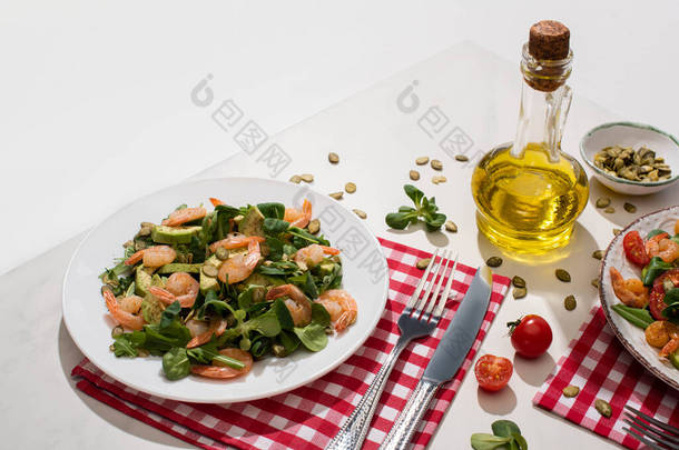 新鲜的<strong>绿色</strong>沙拉，虾仁和鳄梨放在盘子里，靠近餐具，铺在格子布餐巾上，配料放在白桌上