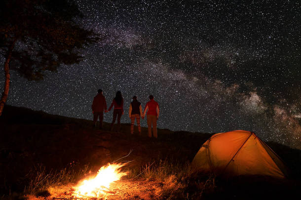 两对徒步旅行者抱着手, 在星空下, 在山上的篝火和橙色帐篷附近露营的夜晚, 在星光灿烂的天空中享受银河。浪漫的夜晚, 在神奇的星空下