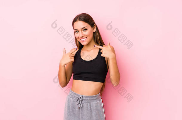 年轻的高加索健身女子穿着<strong>粉色背景</strong>的衣服，微笑着，用手指捂住嘴.
