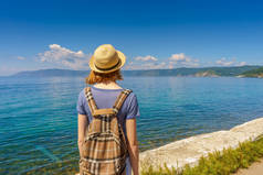 在阳光明媚的夏日里, 来自 listvyanka 村海滨的旅游女孩戴着帽子和背包欣赏贝加尔湖美丽的风景。旅行、自由和暑假概念.