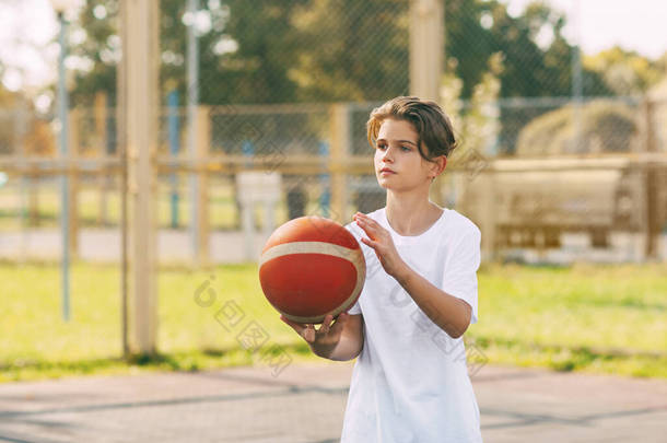 一个穿着白色T恤的漂亮<strong>少年</strong>站在篮球场上，手里拿着一个篮球。体育与健康生活方式的概念