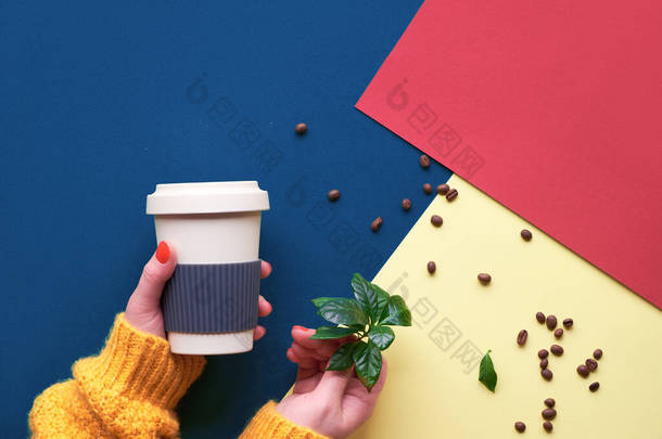 零浪费咖啡的概念。 红色、蓝色和黄色三种色调纸的几何顶视图。环保可重复使用的咖啡杯，手插橙色毛衣，手持杯子和咖啡壶.